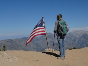 Joanie Kasten stands at Mt. Baden-Powell's summit.  Pine Mountain and Mt. Baldy make up the backdrop as Old Glory flies in a brisk mid-summer breeze.  